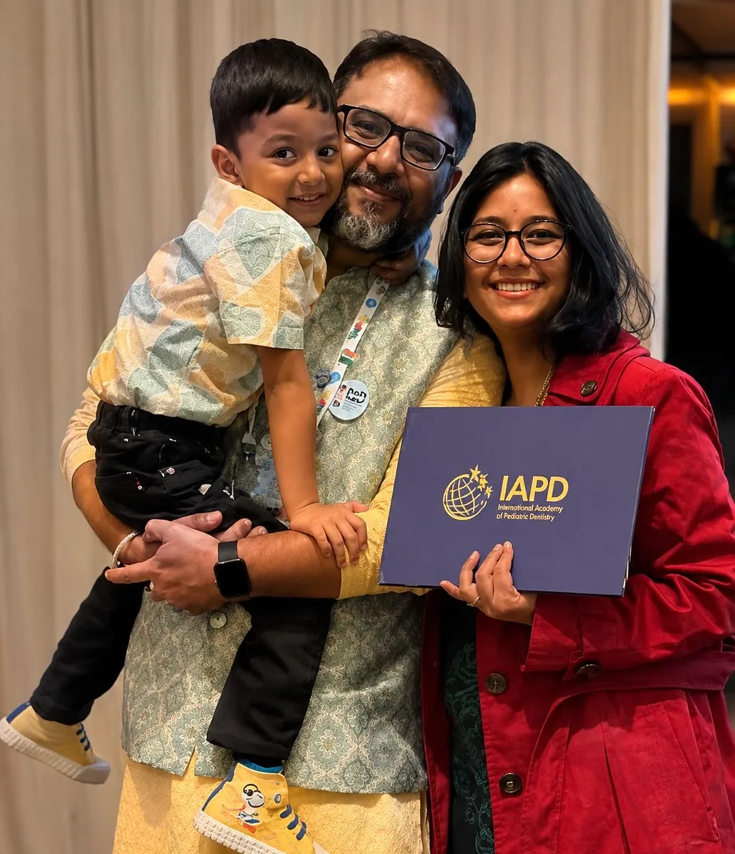 Dr. Sanchit Paul holding a child, beside a woman holding an International Academy of Pediatric Dentistry folder