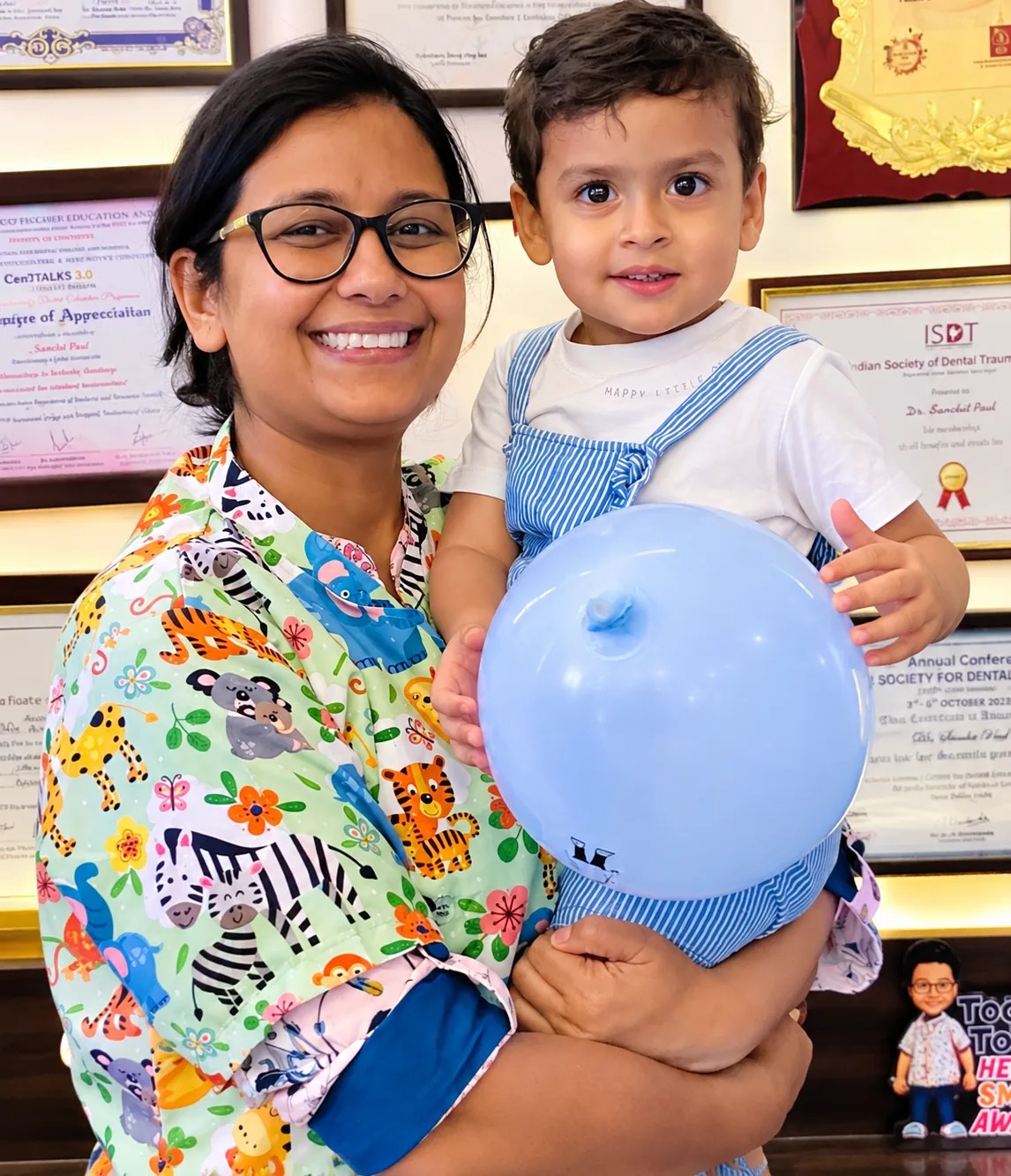 Dr. Shivika Agarwal holding a toddler with a blue balloon, in front of a wall of certificates