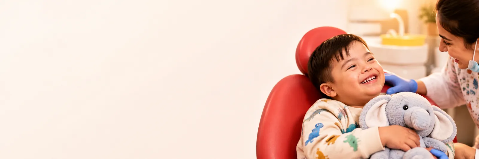 A smiling boy holding a toy elephant in the dental chair while the dentist gently checks his teeth