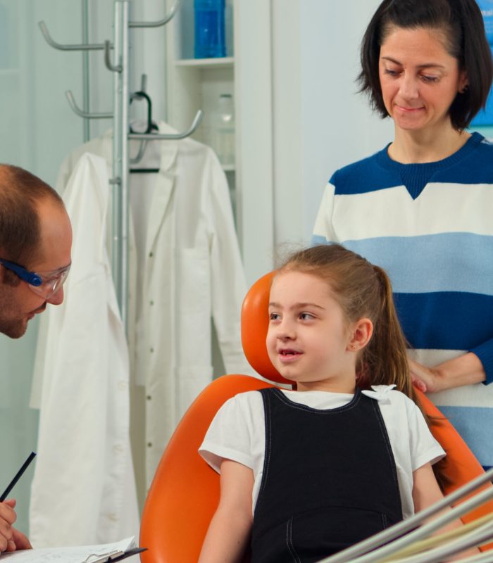 Friendly man dentist assistant interrogating kid patient and taking notes on clipboard waiting for doctor. Mother of little girl explaining dental problem to nurse indicating at toothache problems.